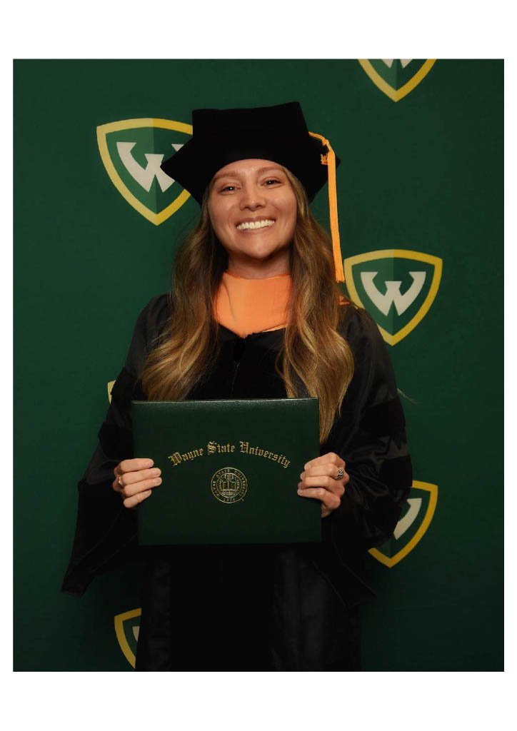 College of Nursing DNP graduate Samantha Sobie stands in front of a green Wayne State Warrior shield backdrop and holding her degree, wearing a black graduation gown and gold doctoral graduation regalia.