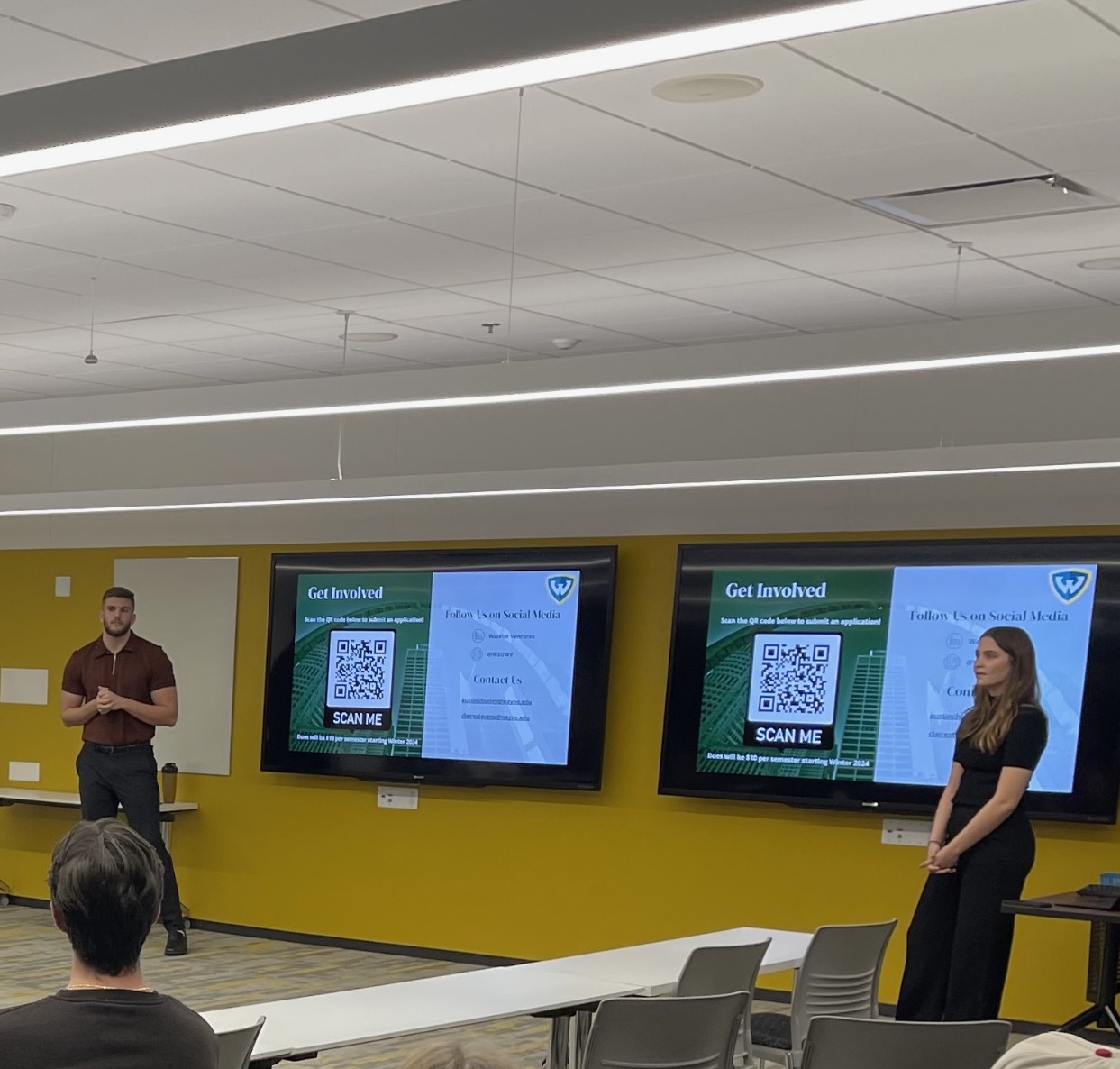 Man and woman stand in front of a screen presenting.