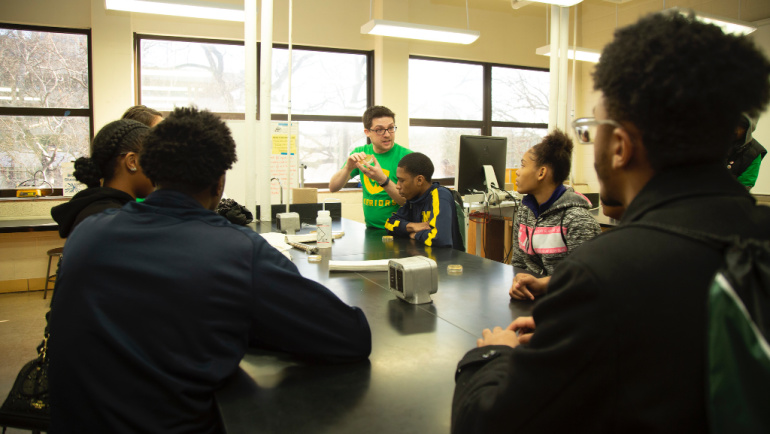 Students gather around a desk, while a Wayne State student demonstrates an experiment.