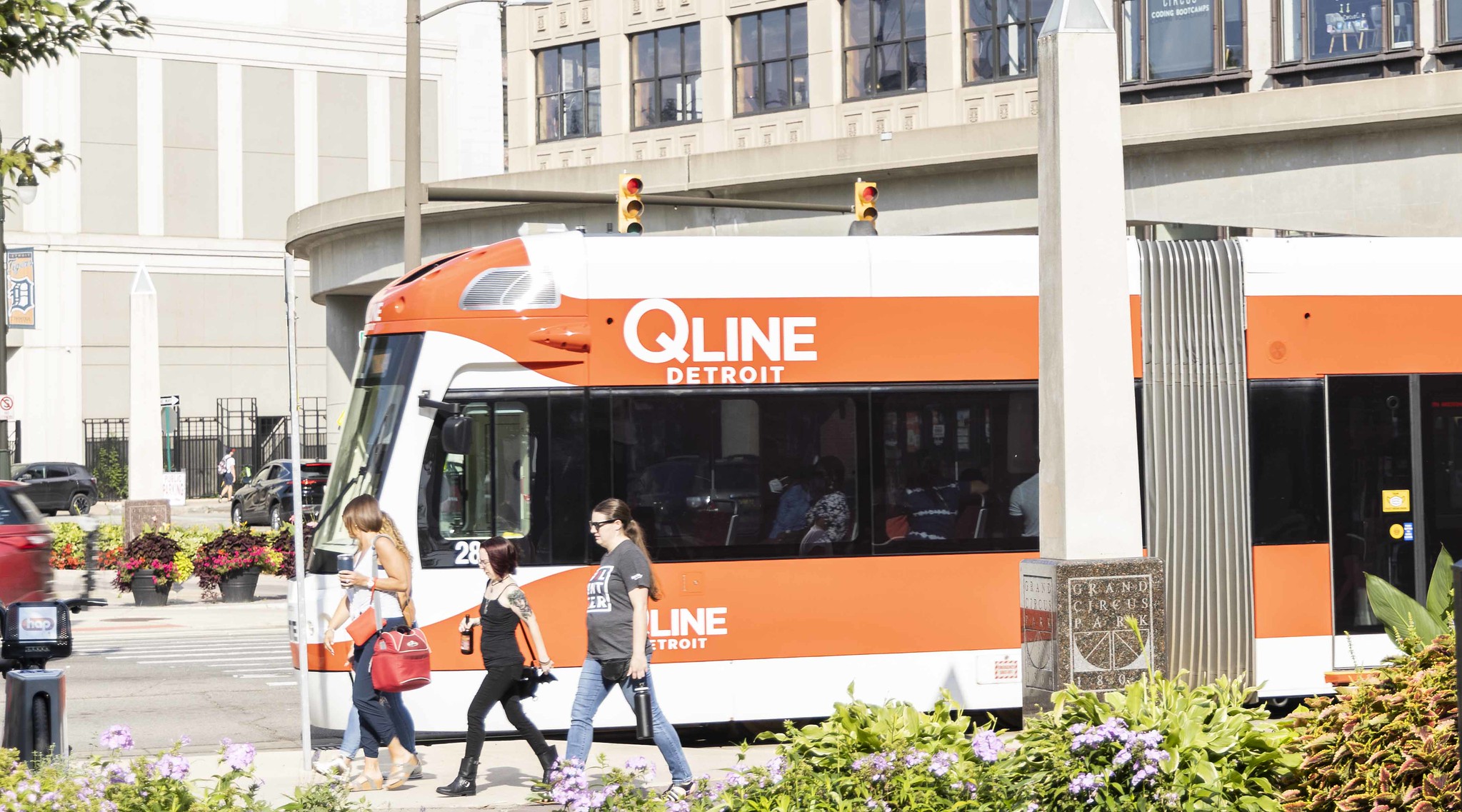 An image of the QLINE street car driving on Woodward with people walking on the sidewalk to the stop.