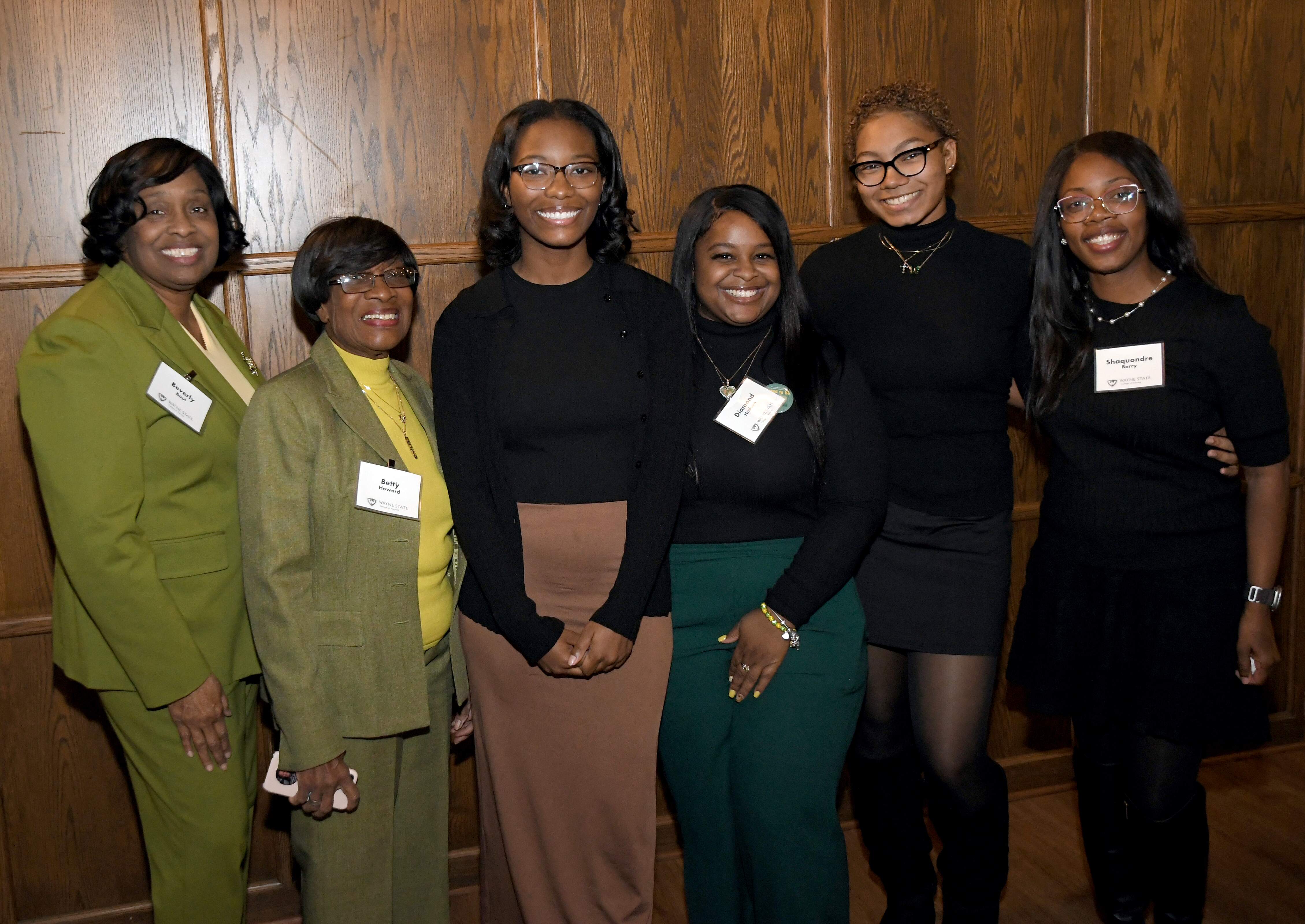 A group of six women stand next to each other with their arms around each other for a photo, four people are wearing black and two people are wearing lime green to the left.