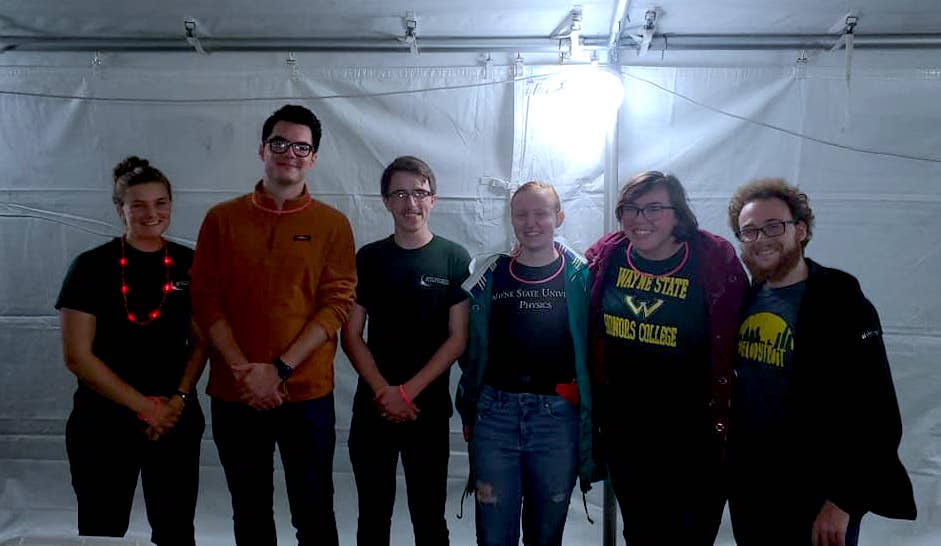 Six people stand in a row smiling, inside of a white tent.  Five of them are wearing Wayne State shirts.