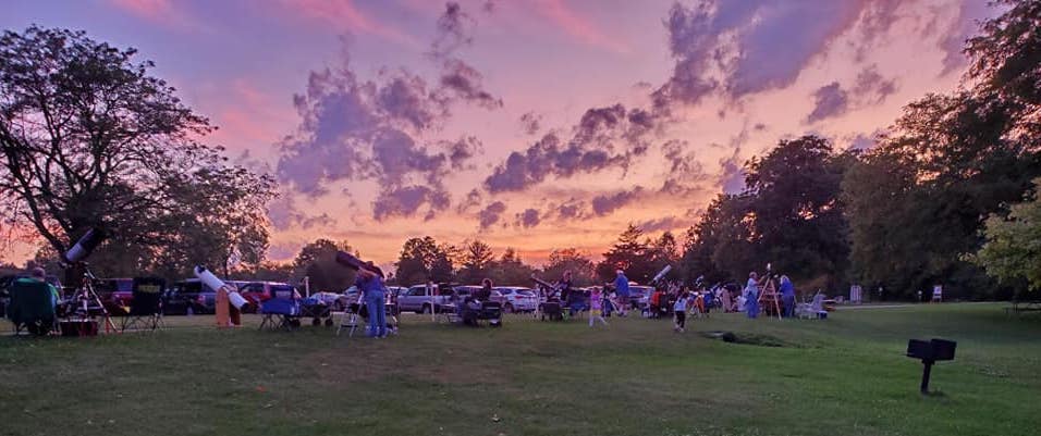People sitting on grass lawn with telescopes while the sunset behind them is orange and purple.