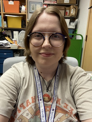 SIS student Amber Carnahan is pictured in front of shelves in a storage room. She has short medium brown hair and is wearing round frame glasses and a slight smile. 