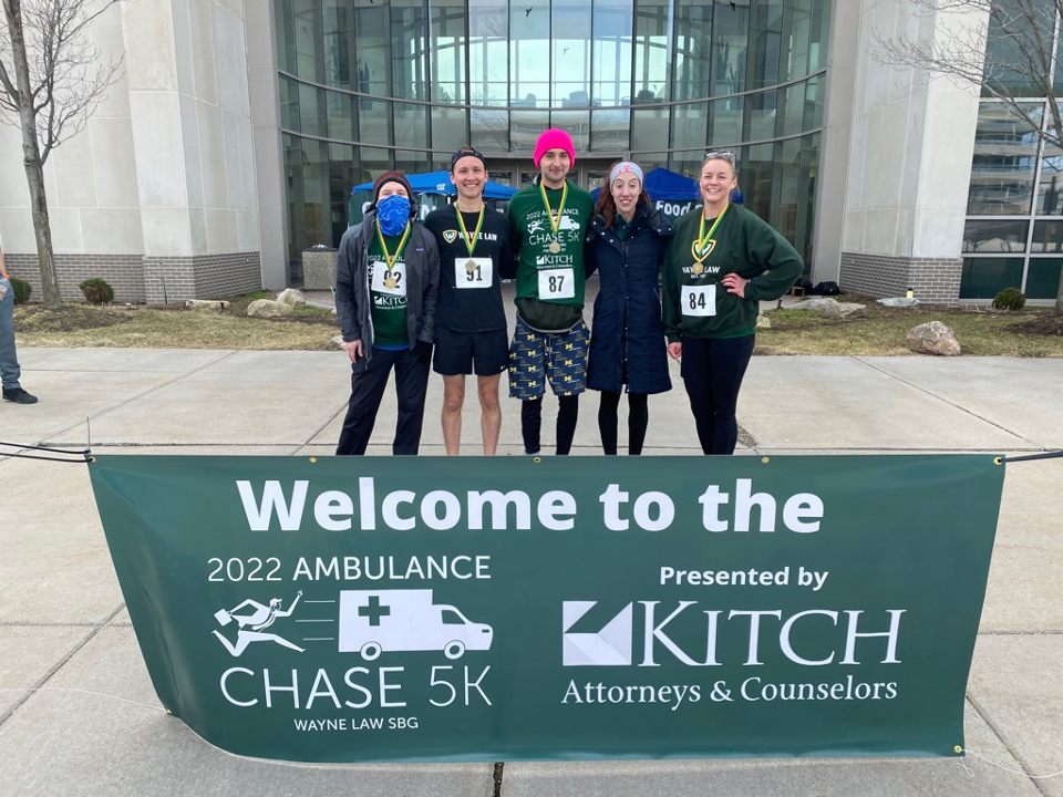 Runners pose in front of Wayne State University Law School building in front of a sign that reads, Welcome to the 2022 Ambulance Chase 5K presented by Kitch Attorneys & Counselors