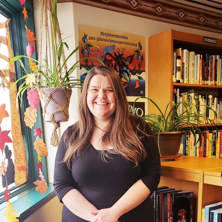 School of Information Sciences student Angela Badke is pictured in a library in front of reading posters and bookshelves. She has long light brown hair and a warm smile. 