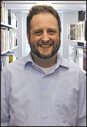 SIS alumnus Calvin Battles is pictured in front of a tall library bookshelf. He has short hair and facial hair and is smiling at the camera.