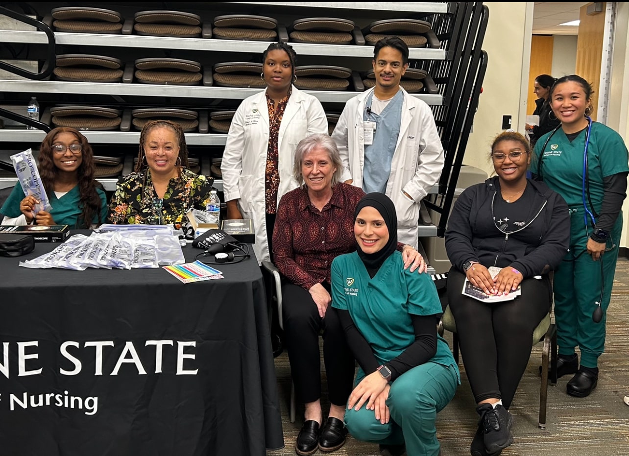  A group of eight College of Nursing students and faculty stand next to a table with a Wayne State College of Nursing tablecloth. Three people are wearing blue-green scrubs and two people are wearing white coats.