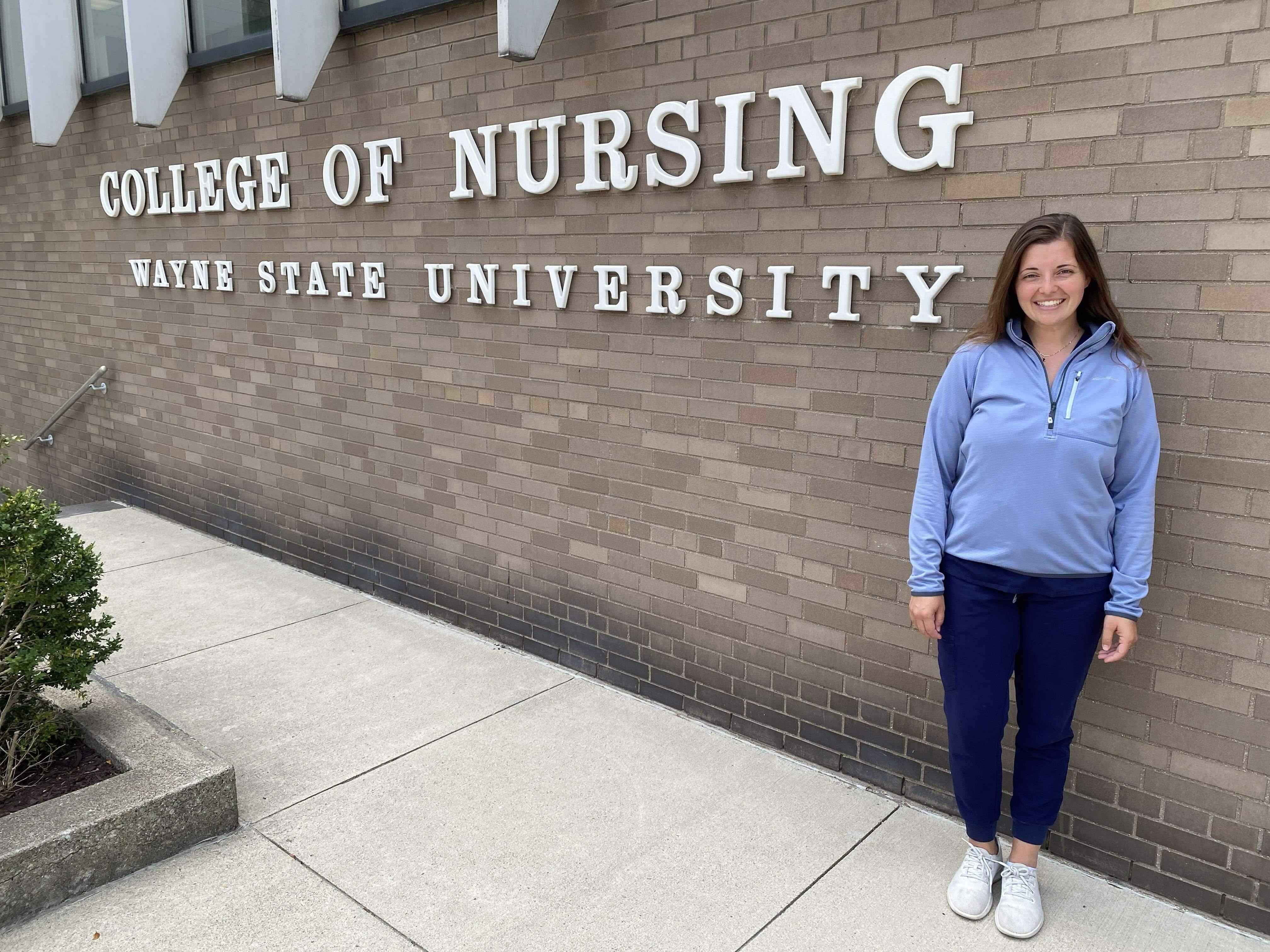 Nursing student Annika Bajema stands in front of the grey brick Wayne State College of Nursing building, wearing a light blue sweater and her brown hair on her shoulders.