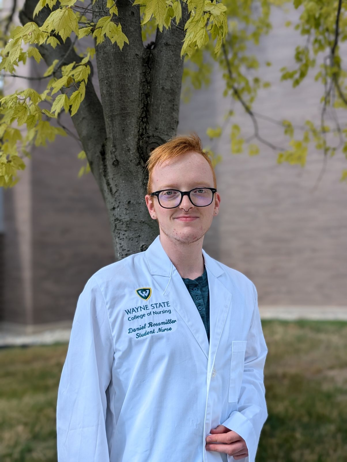 Nursing student Daniel Ross Miller stands in front of a tree, wearing his College of Nursing white coat smiling at the camera with his ginger short hair and black glasses.