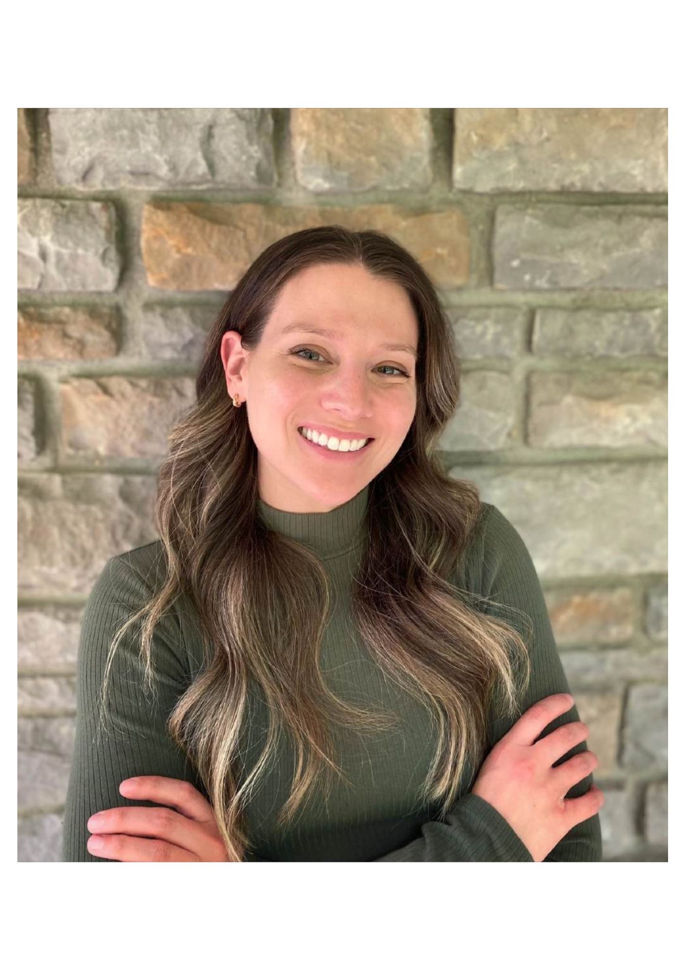 College of Nursing DNP graduate Samantha Sobie stands in front of a grey brick wall with her arms crossed and smiles for a photo, wearing a green turtle neck sweater and her dark blond hair down on her shoulders.