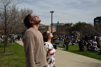 Two observers wearing solar eclipse glasses staring at the Sun. A multitude of other observers are seen in the background.