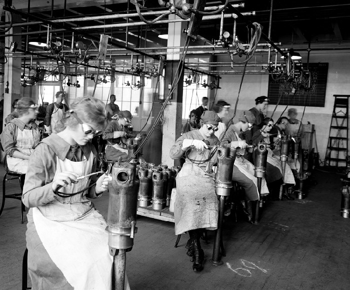 A large team of women wearing protective goggles and aprons and working in a factory with torches