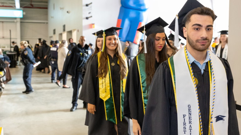 A group of graduates walk during the traditional commencement march, a few proudly displaying 'First Generation' stolls on their graduation regalia.