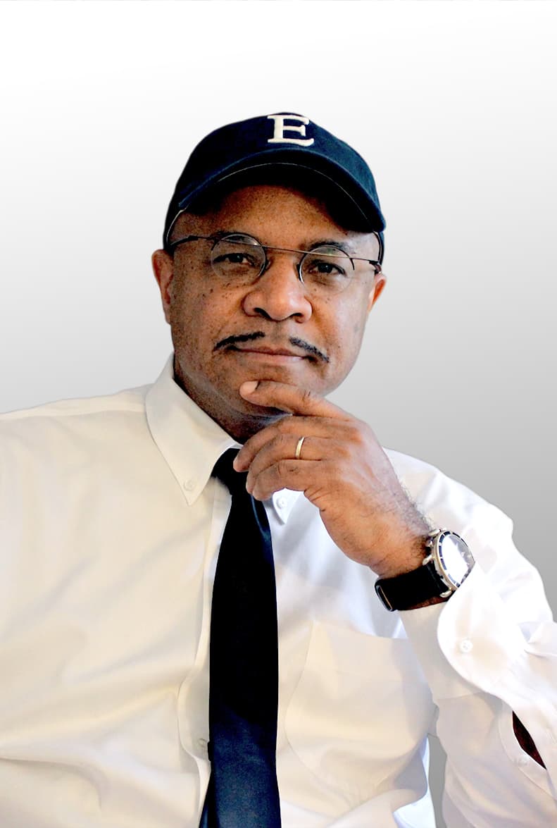 A man in a white shirt, dark tie, glasses and a baseball cap in front of a white background.