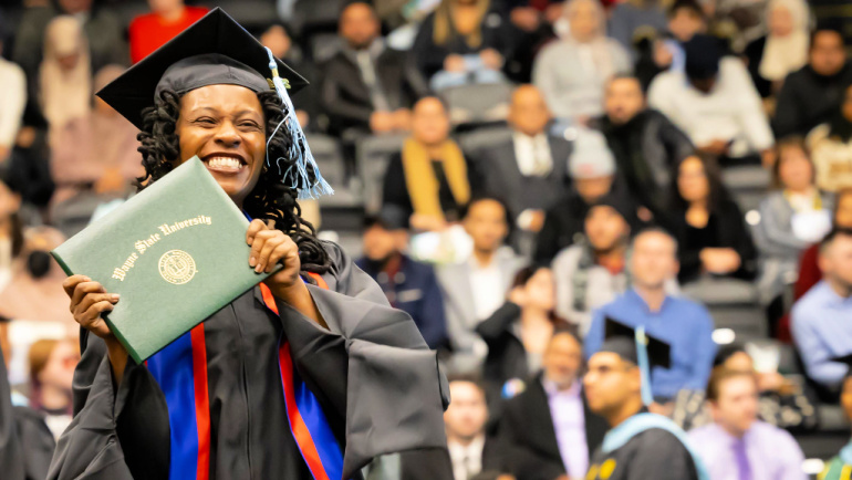 A graduate, wearing a cap and gown at commencement, holds her Wayne State diploma, grinning from ear to ear.