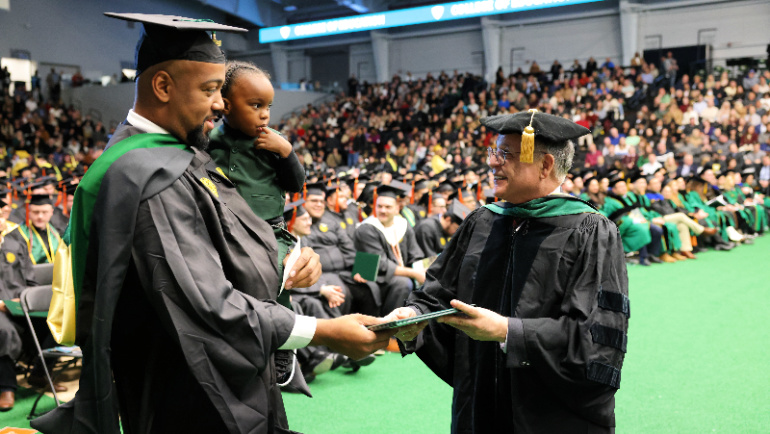 A smiling graduate accepts his master's degree while holding his young son.