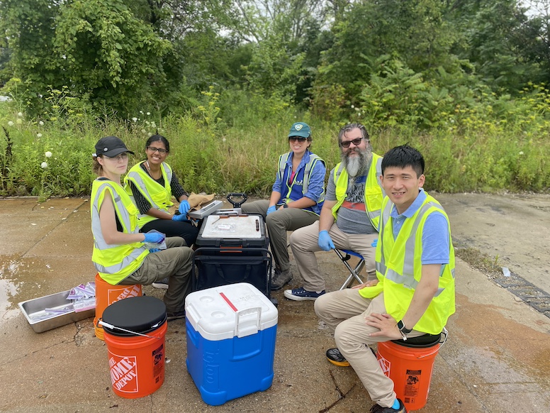 People in neon vests sit atop orange buckets by coolers and equipment on a paved surface by a forested area