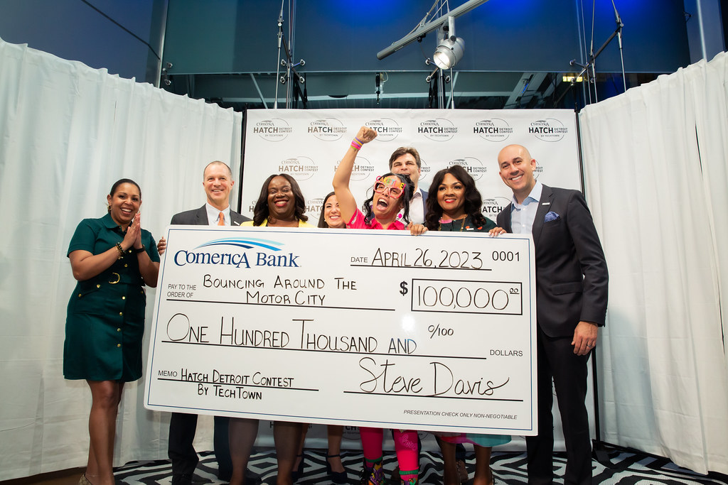 A woman triumphantly shouts with her fist in the air as she holds a large check for $100,000 from Comerica Bank for her business, Bouncing Around The Motor City. Representatives from Comerica Bank and TechTown Detroit smile and pose beside her