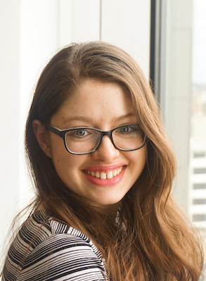 A head and shoulders photo of Frances Heldt, a young Caucasian woman. She is turned to look over her shoulder. She has long chestnut colored hair and is wearing glasses, a black and white stripped top and a big smile.