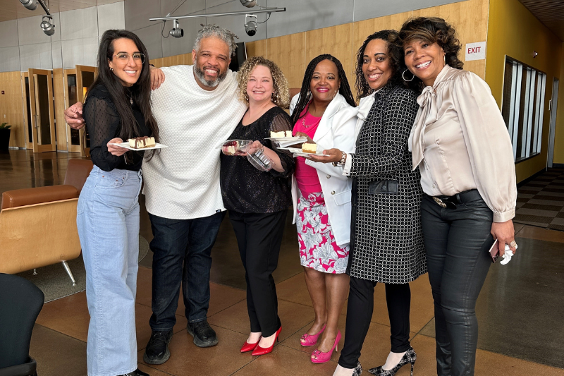 A group of six people smile and pose for a photo. Two of them are holding plates with dessert on them.