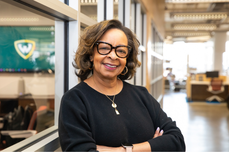 A photo of a woman smiling with her arms crossed in front of her. She is standing in a coworking office.