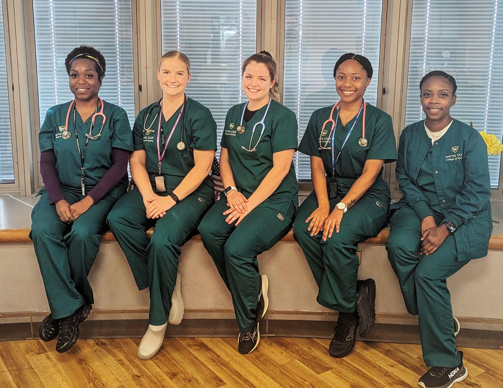 A group of four women sit together in front of windows for a photo and they all are wearing blue green scrubs.