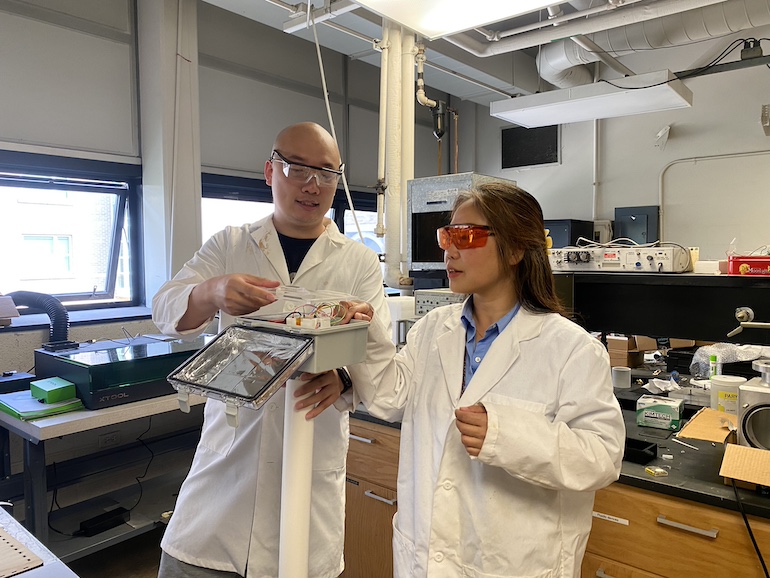 Dr. Wager and her student Qisen are in middle of photo in white lab coats and safety glasses. They are both holding and looking at their sensor.