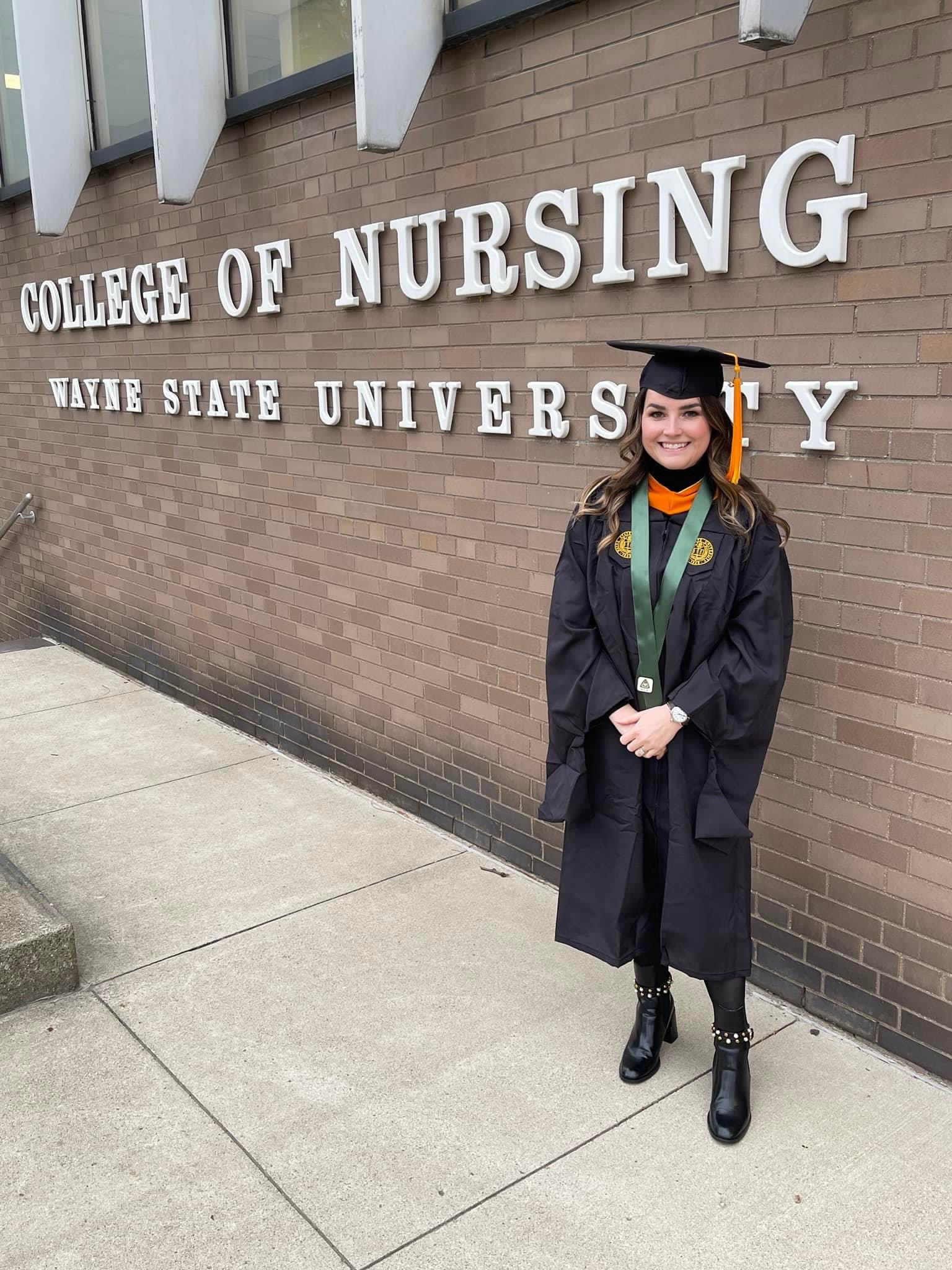 Kaitlin Bratby stands in front of a gray brick wall with white letters on it that spell out Wayne State University College of Nursing wearing a black graduation cap and gown with a gold tassel.