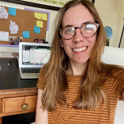 Amanda Kemp, a Caucasian woman with long, light brown hair and glasses is pictured taking a selfie in front of her home workstation. She is wearing a rust colored short sleeve top and a smile.