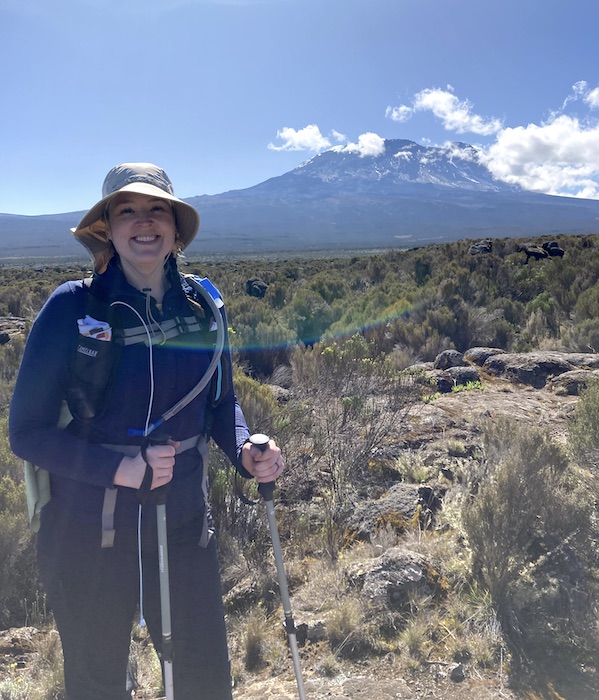 Emily is pictured in front of Mount Kilimanjaro. She is wearing a wide-brimmed hat and is carrying walking sticks to aid her during the climb.
