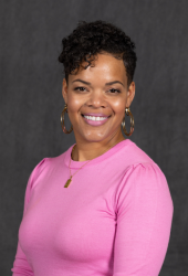 A headshot of SIS Professor Kumasi. She is wearing a vibrant pink top and gold hoop earrings. She has short, curly brown hair.