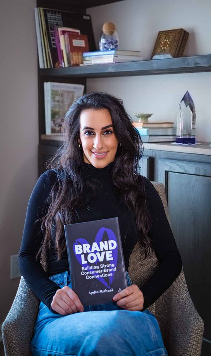 Wayne State Ilitch School alumna Lydia Michael sitting in a chair holding her debut novel, Brand Love.