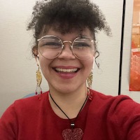 Madelynne is wearing large, gold-rimmed glasses and gold dangly earrings. She has curly brown bangs and a friendly smile.