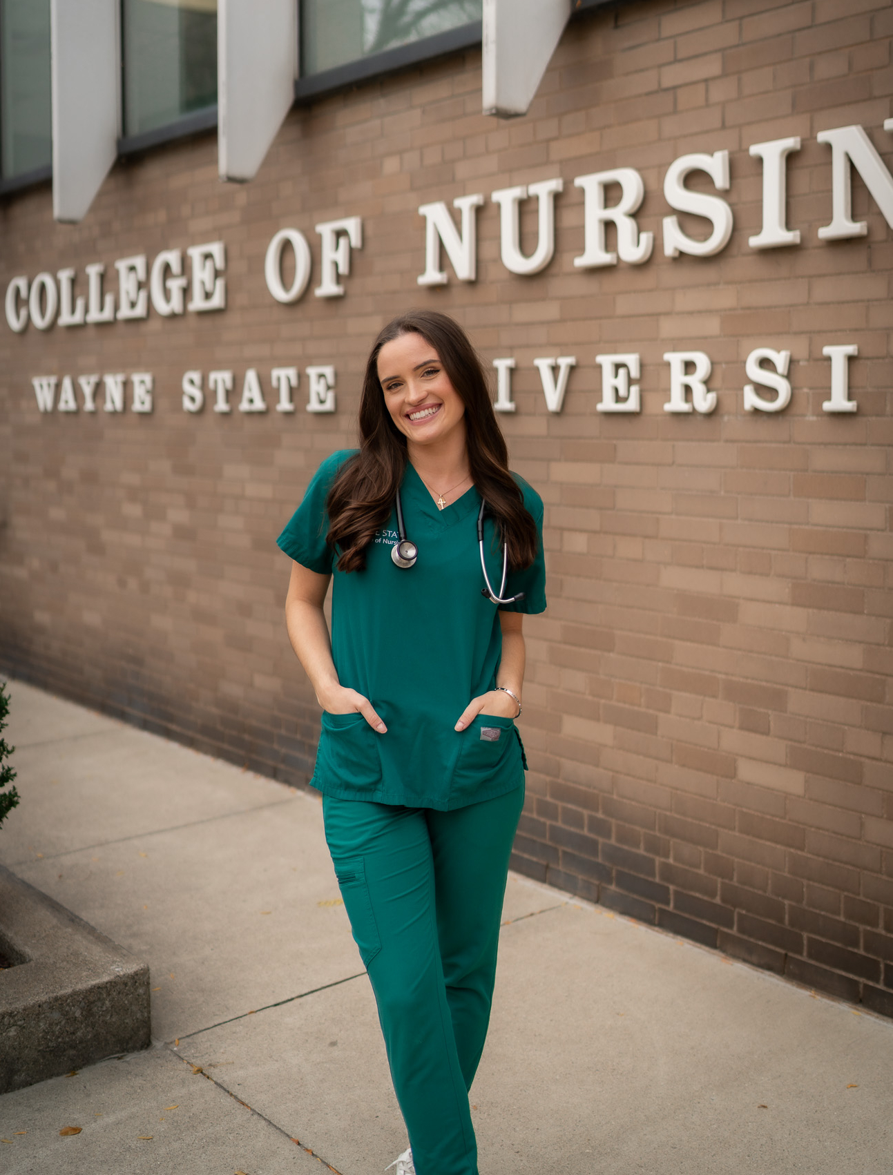 College of Nursing graduate Mary Forbes stands in front of the grey brick Wayne State University College of Nursing building wearing blue-green scrubs and a black stethoscope over her shoulders.