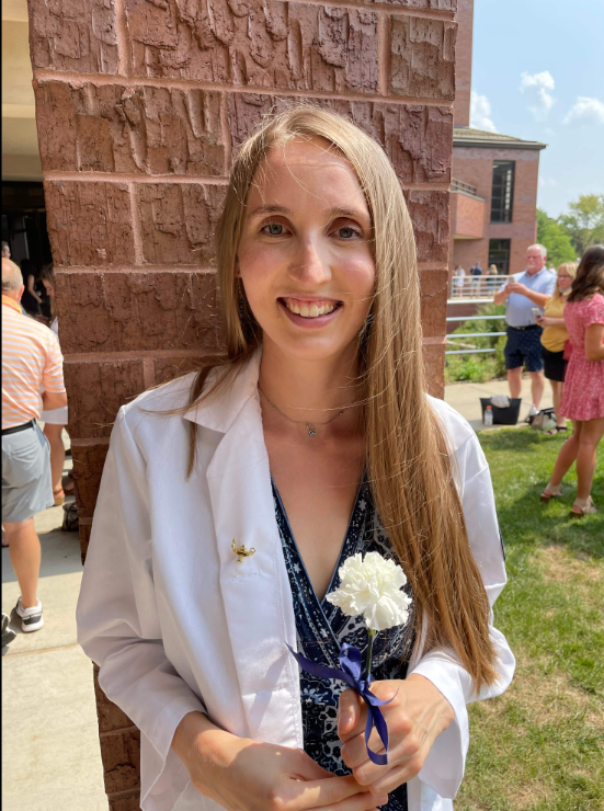 Nursing student Melanie Hampton smiles at the camera wearing a white coat, standing in front of orange brown brick wall, holding a white flower in her hands.