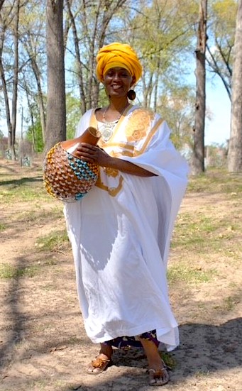  Michelle Jahra McKinney is a Black woman dressed in a traditional African kaftan dress. It is white with decorative yellow trim around the neck. She wears a matching yellow head scarf and holds a beaded jug instrument.  