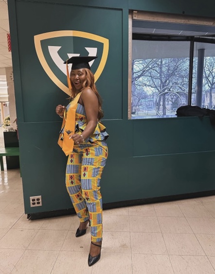 BSN graduate Nennie Sheriff standing, wearing a yellow and blue square patterned pant suit with a black graduation cap on her head and gold stole over her shoulders.