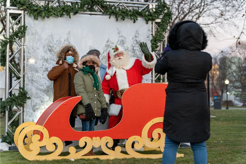  Three people stand next to an individual dressed as Santa Claus and pose for a photograph. They stand in front of a snowy backdrop and behind a red sleigh. An individual is in front of the people and scenery taking a photograph