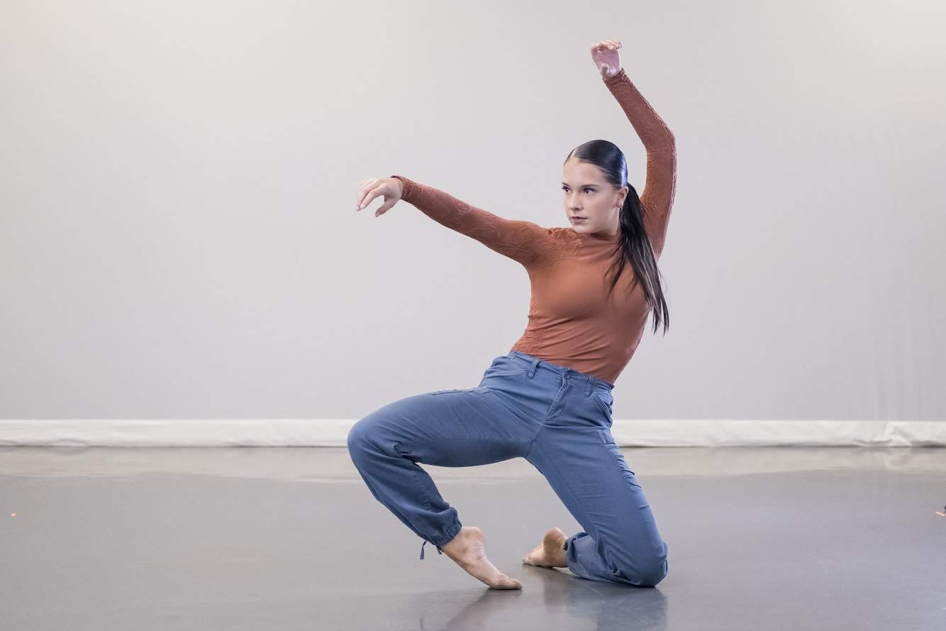  Senior dance major Mallory Marshall is captured dancing in front of a white backdrop wearing an orange long sleeve shirt and blue pants