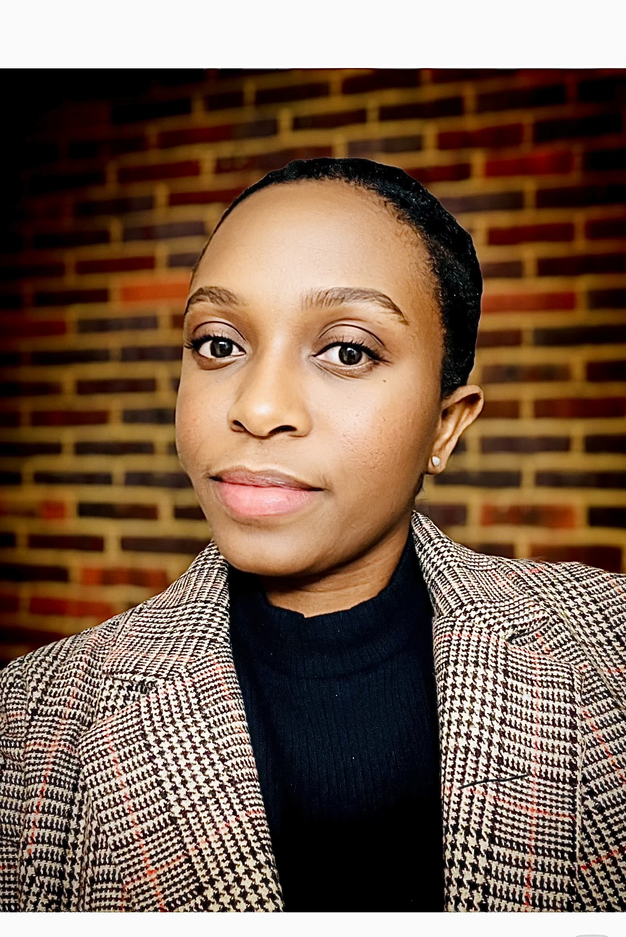 Nursing student Rachel Anyu Gyesbour poses for a portrait photo in front of a brown brick wall, wearing a black shirt underneath a light brown striped blazer.