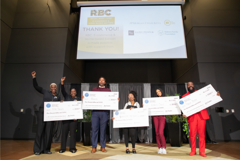 Six people stand on a stage in an auditorium, holding large checks from TechTown Detroit for their respective businesses.