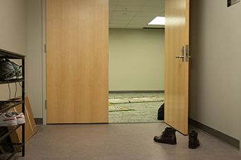 Two wooden double doors reveal one of the entrances to the renovated Student Center Reflection Room at Wayne State. A pair of brown boots sit in front of the door. To the left of the door sits a shoe rack with two pairs of shoes on it.