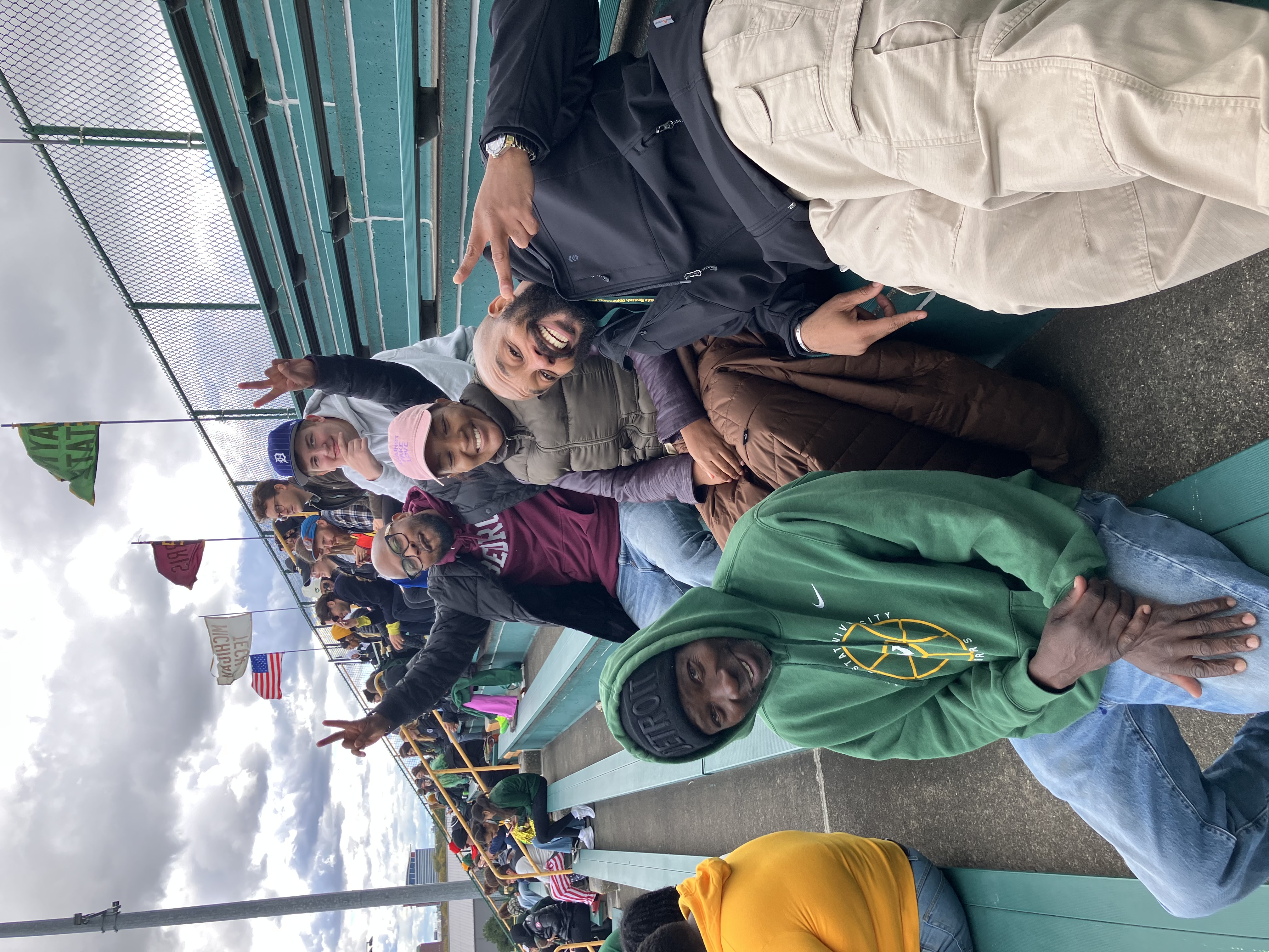 Five students posing on a set of bleachers with other seated spectators