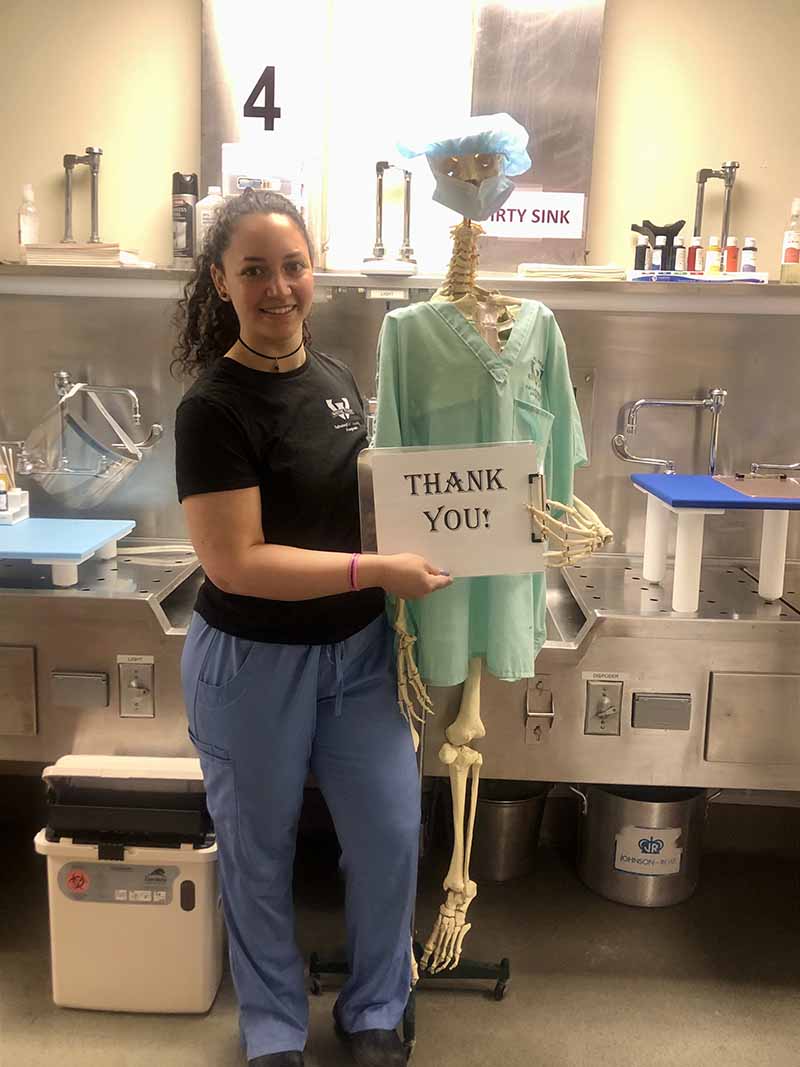 Wayne State pathologists' assistant student, wearing blue scrub pants, a black Wayne State shirt, standing next to a skeleton model wearing a blue scrub cap