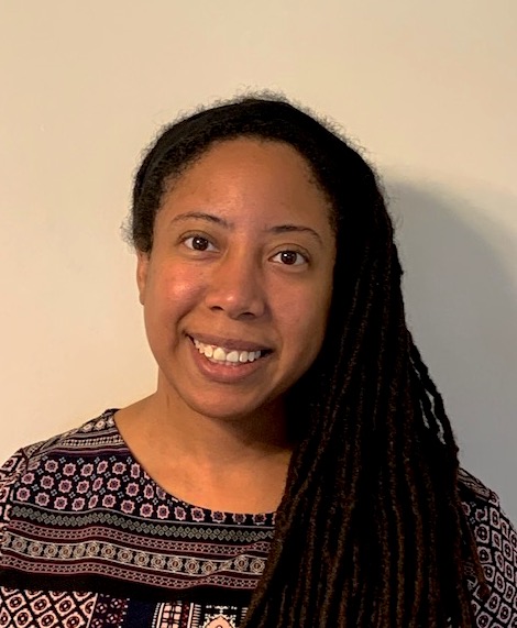 Sarah is a Black woman with long dark hair that is twisted into spirals. She is wearing a printed top and is smiling at the camera.