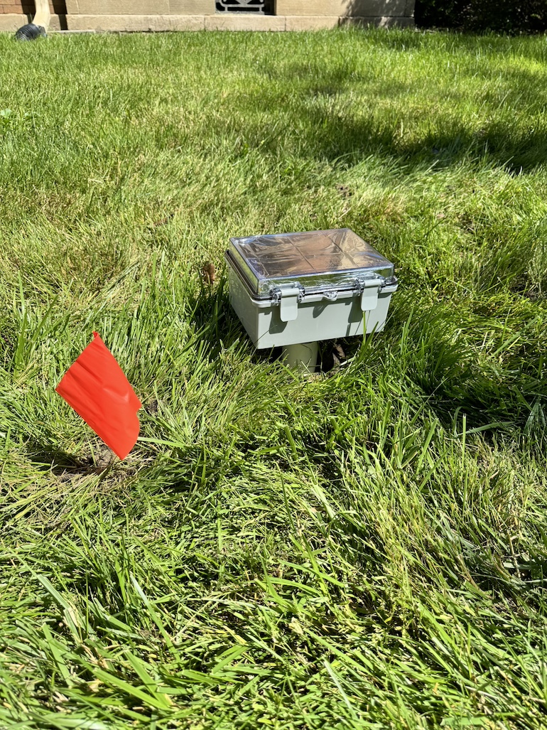 The sensor prototype (a light colored box with lid) sits on top of a white pipe in green grass. Orange flag on the left to signal device presence.