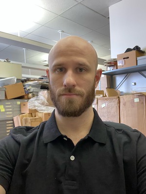 SIS student Spencer Miller is pictured in front of an archive storage room. He has a bald shaved head and medium brown facial hair. He is wearing a black polo shirt and a serious expression.