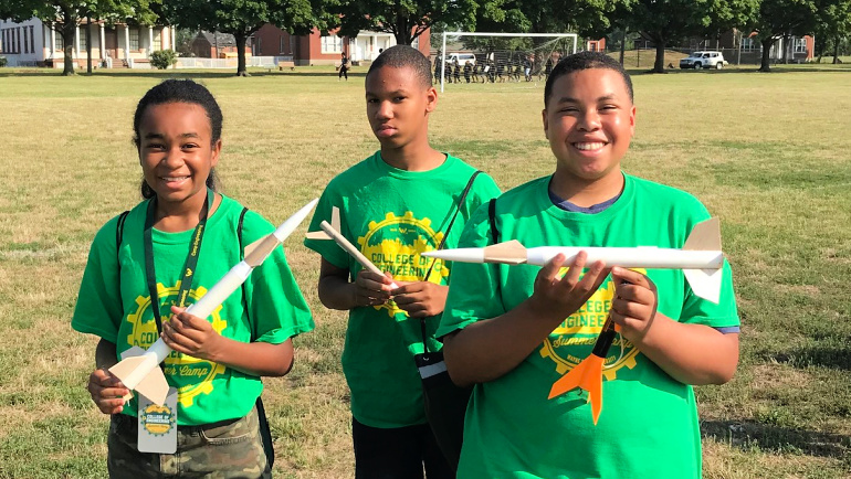 Young students stand outside with bottle rockets they have built in summer camp.