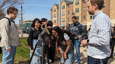 A student peers through a telescope while behind them, a line of other students awaits their turn for observation. Overseeing the activity is Dr. Gonderinger, positioned on the right.
